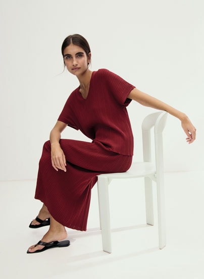 Look 6 entdecken: Our Favourite New Looks: Frau im bordeauxfarbenen Strickensemble, modern auf hellem Stuhl im Studio inszeniert.