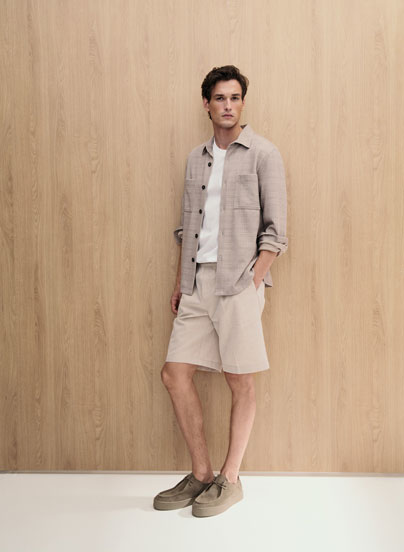 Look 4 entdecken: Our Favourite New Looks: Mann im hellen Sommer Set aus offener Hemdjacke, Shirt und Shorts, lässig vor warmer Holzfläche inszeniert.