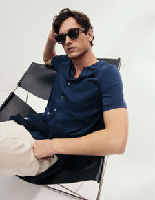 Look 1 entdecken: Our Favourite New Looks: Mann im navyfarbenen Stricktop und heller Hose, locker im Designerstuhl positioniert, minimalistisches Studio Setting.