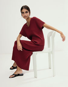 Look 6 entdecken: Our Favourite New Looks: Frau im bordeauxfarbenen Strickensemble, modern auf hellem Stuhl im Studio inszeniert.