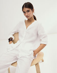 Look 3 entdecken: Our Favourite New Looks: Frau in lockerem weißem Leinen Set aus Bluse und Hose, minimalistisch im Studio auf einem hellen Stuhl inszeniert.