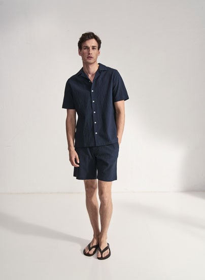 Our Favourite New Looks: Mann im navyfarbenen Sommer Set aus Kurzarmhemd und Shorts, clean im Studio präsentiert – lässiger Look zum Nachstylen.