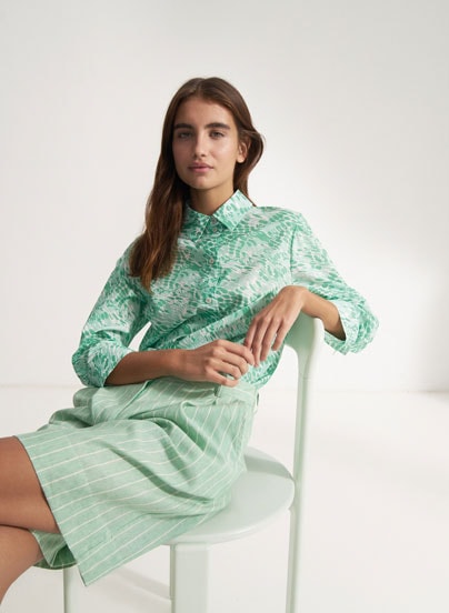 Our Favourite New Looks: Frau in gemusterter, mintfarbenen Sommerbluse und gestreifter Shorts, entspannt auf Stuhl inszeniert – moderner Premium Look zum Nachstylen.