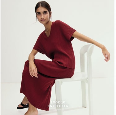 Look 10 entdecken: Our Favourite New Looks: Frau im bordeauxfarbenen Strickensemble, modern auf hellem Stuhl im Studio inszeniert.