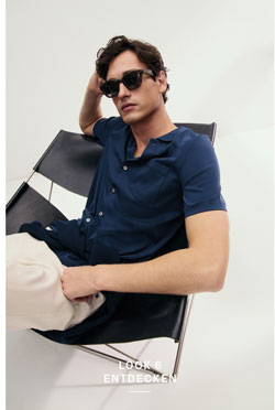 Look 6 entdecken: Our Favourite New Looks: Mann im navyfarbenen Stricktop und heller Hose, locker im Designerstuhl positioniert, minimalistisches Studio Setting.