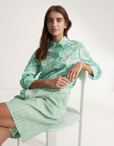 Our Favourite New Looks: Frau in gemusterter, mintfarbenen Sommerbluse und gestreifter Shorts, entspannt auf Stuhl inszeniert – moderner Premium Look zum Nachstylen.