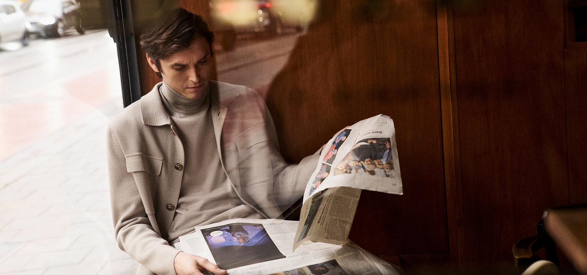 MONDAY TO FRIDAY: Mann im hellen Strick Tailoring mit Rollkragen und Cardigan, beim Zeitunglesen im urbanen Café Setting präsentiert, moderne Premium Workwear.