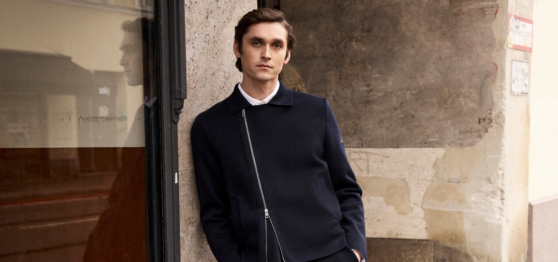 MONDAY TO FRIDAY: Mann in navyfarbener Strickjacke mit Reißverschluss, lässig vor urbaner Architektur präsentiert, zeitlose Premium‑Workwear.