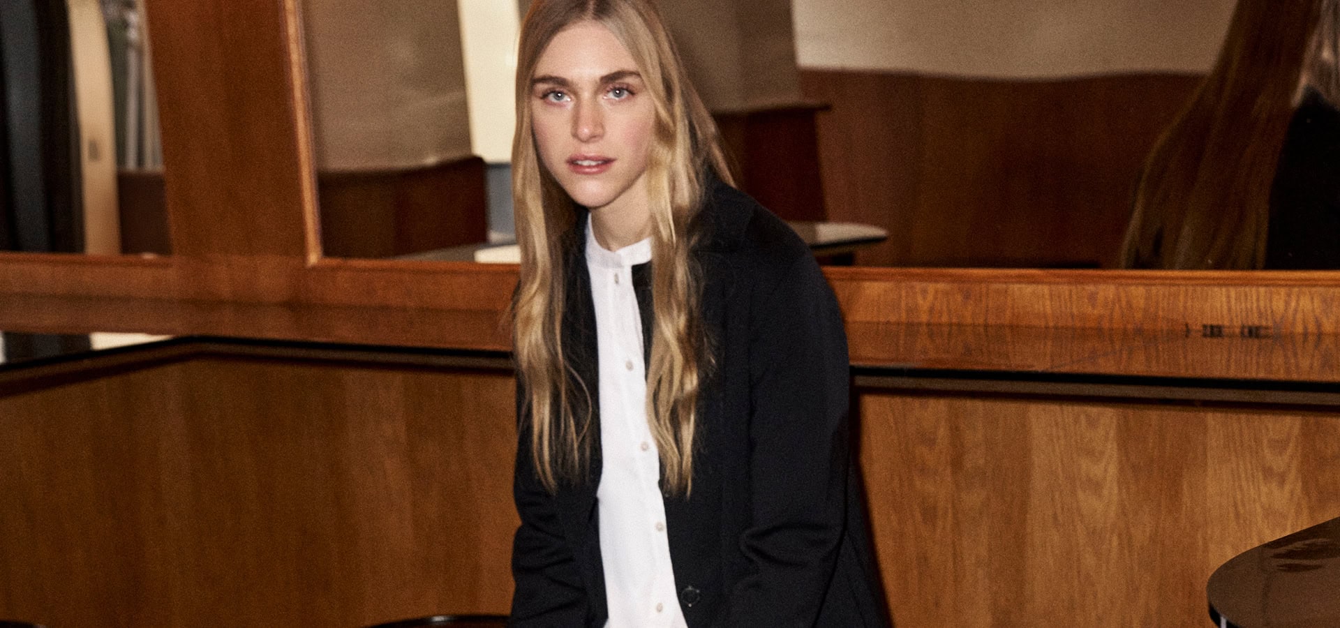 MONDAY TO FRIDAY: Frau im dunklen Strickblazer über heller Bluse, modern im Interieur mit warmer Holzvertäfelung inszeniert.