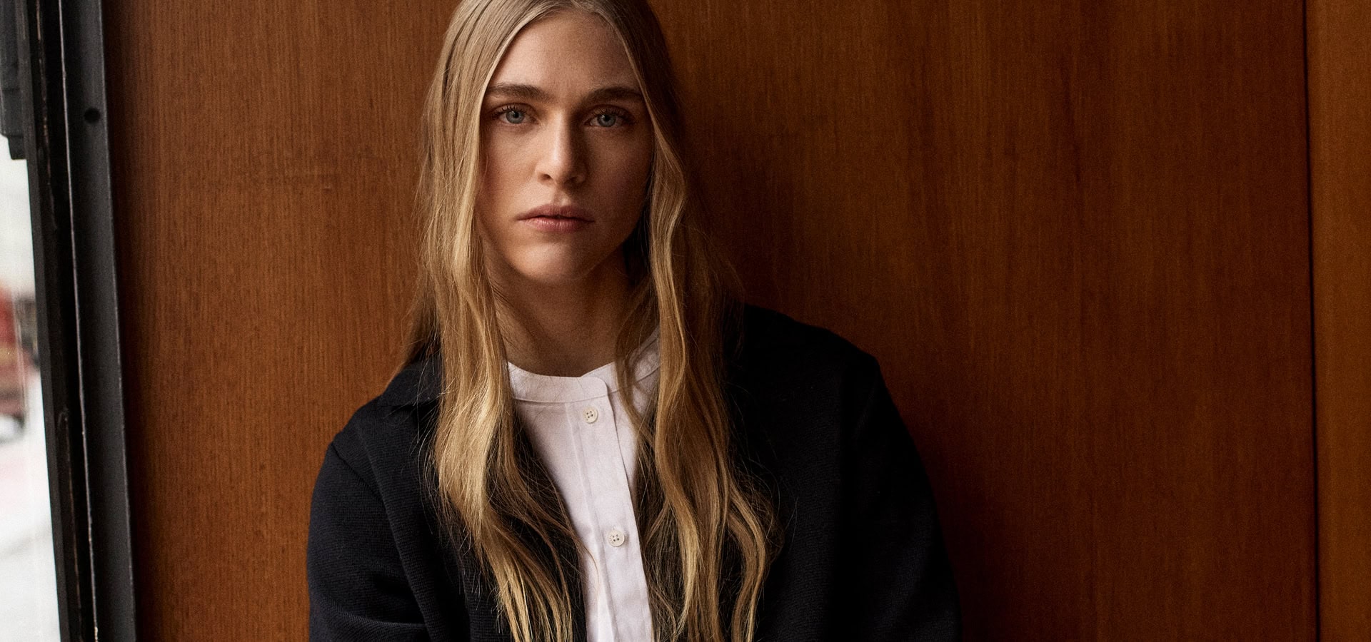 MONDAY TO FRIDAY: Frau im dunkelblauen Business‑Casual Look mit Strickjacke und hellem Top, stilvoll vor warmer Holzvertäfelung präsentiert.