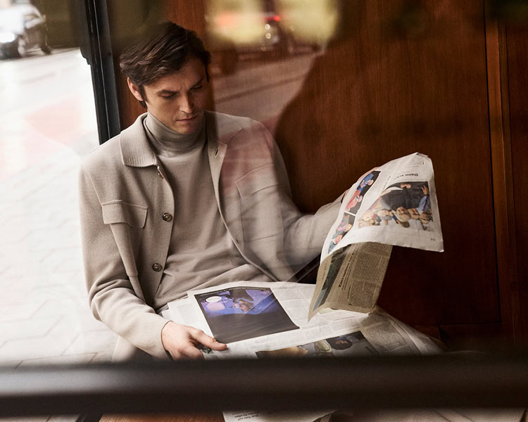 MONDAY TO FRIDAY: Mann im hellen Strick Tailoring mit Rollkragen und Cardigan, beim Zeitunglesen im urbanen Café Setting präsentiert, moderne Premium Workwear.