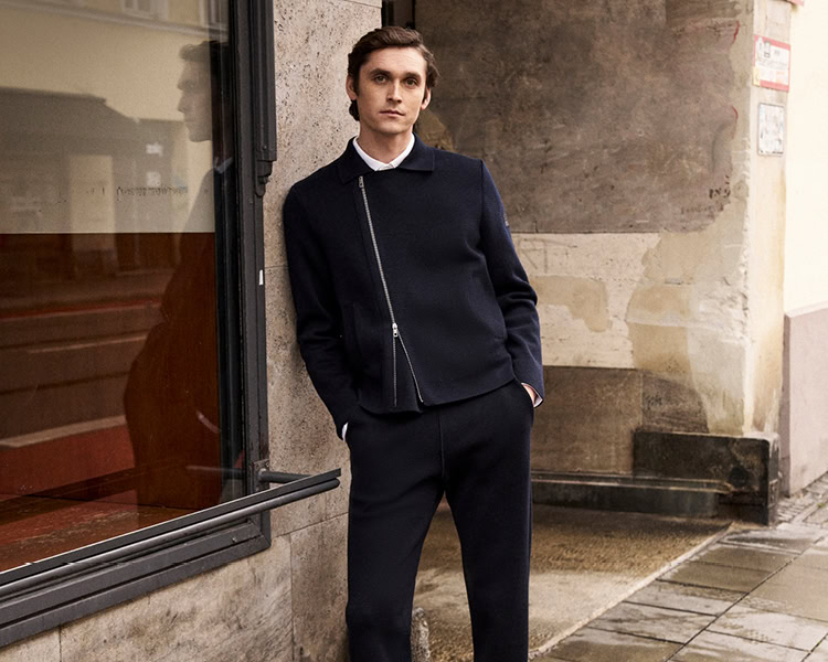 MONDAY TO FRIDAY: Mann in navyfarbener Strickjacke mit Reißverschluss, lässig vor urbaner Architektur präsentiert, zeitlose Premium‑Workwear.