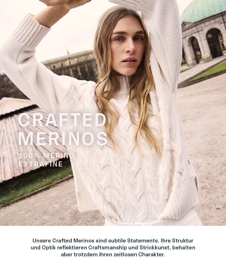 CRAFTED MERINO CRAFTED MERINOS Women: Frau im hellen Zopfstrickpullover aus 100 % Merino extrafine vor historischer Architektur präsentiert. Strukturierte Merino‑Styles mit zeitloser Strickkunst und hochwertiger Verarbeitung.
