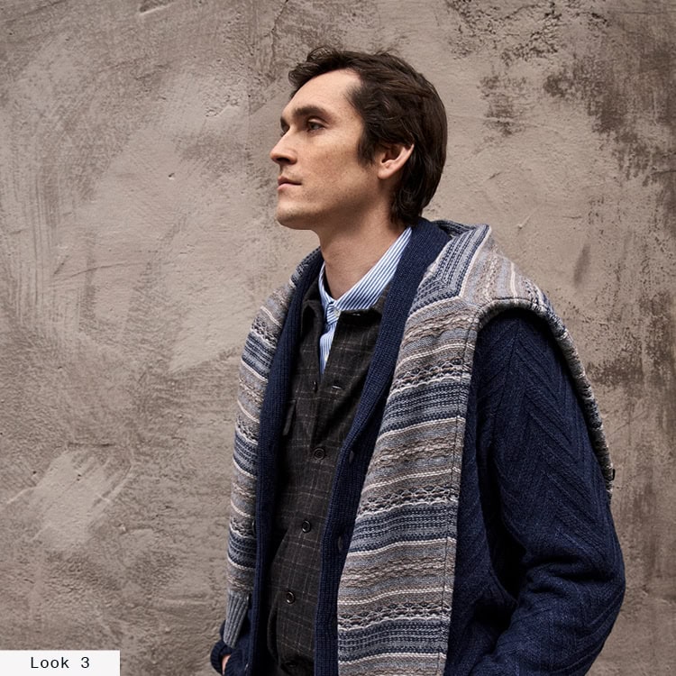 Look 6 English Formality Men: Mann in navyfarbener Strickjacke über Hemd und Feinstrickpullover mit gemustertem Pullover über den Schultern vor strukturierter Steinwand.