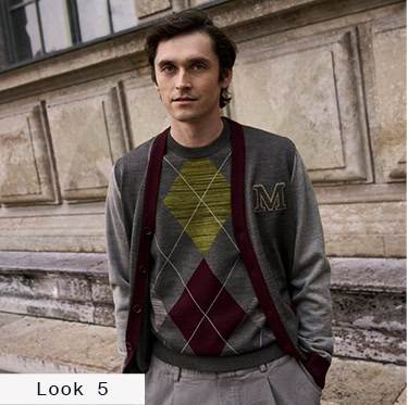 Look 5 English Formality Men: Mann im Strickpullover mit Rautenmuster unter einer Strickjacke mit Kontrastdetails und Stoffhose vor historischer Architektur.