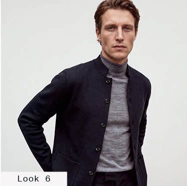 Look 6 English Formality Men: Mann in navyfarbener Strickjacke über grauem Rollkragenpullover vor hellem Hintergrund.