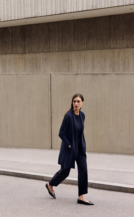 MAERZ Muenchen – Merino Classics Frau in dunkelblauem Strickoutfit mit langer Jacke und passender Hose, unterwegs vor Betonwand.