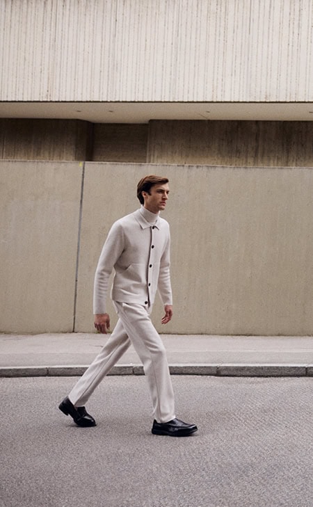 MAERZ Muenchen – Merino Classics Mann geht in hellem Outfit mit Stehkragenjacke und passender Hose vor urbaner Betonwand.