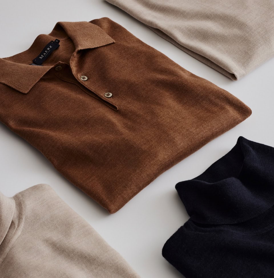 Gefaltete Premium-Merinopullover in Beige, Braun und Dunkelblau, ordentlich arrangiert auf heller Fläche.