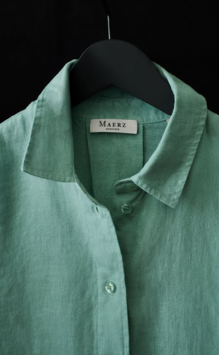 MAERZ Muenchen – Linen Grünes Leinenhemd auf schwarzem Kleiderbügel vor dunklem Hintergrund, sichtbares Label ‚Maerz‘.