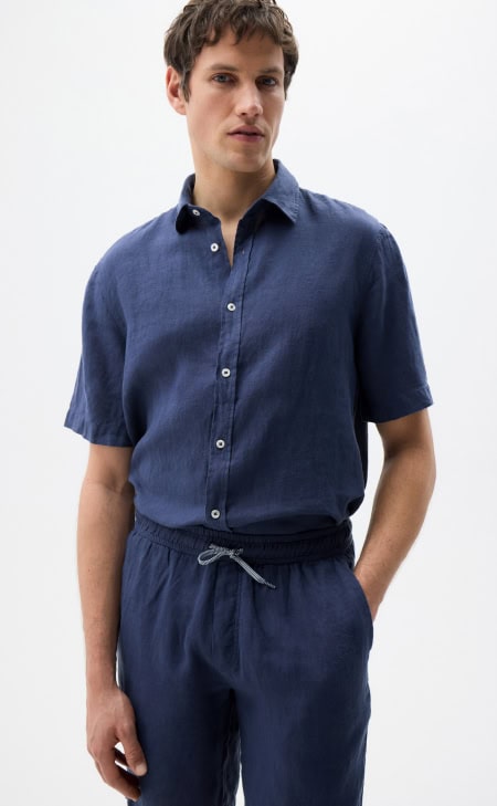 MAERZ Muenchen – Linen Mann trägt dunkelblaues Leinenhemd mit kurzer Knopfleiste und passende Leinenhose, minimalistischer Look.