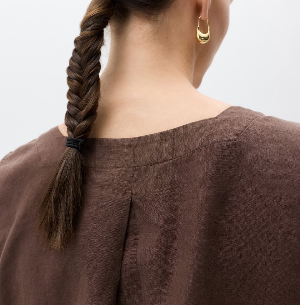 MAERZ Muenchen – Linen Nahaufnahme von hinten: Eine Frau trägt braunes Leinenoberteil mit weitem Ausschnitt, geflochtener Zopf und goldfarbenem Ohrring.