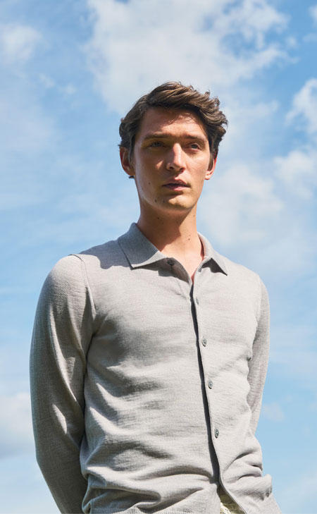 Mann in softem Merino‑Air Poloshirt vor klarem Himmel, modern und ruhig präsentiert – leichter Premium‑Strick für sommerliche Looks.