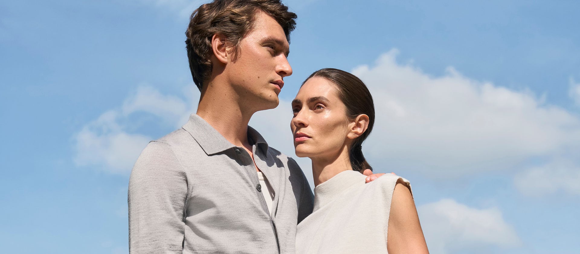 Mann und Frau in leichten Merino‑Air Looks vor blauem Himmel, modern und klar inszeniert – extrafeiner Strick für luftige Premium‑Sommermode.