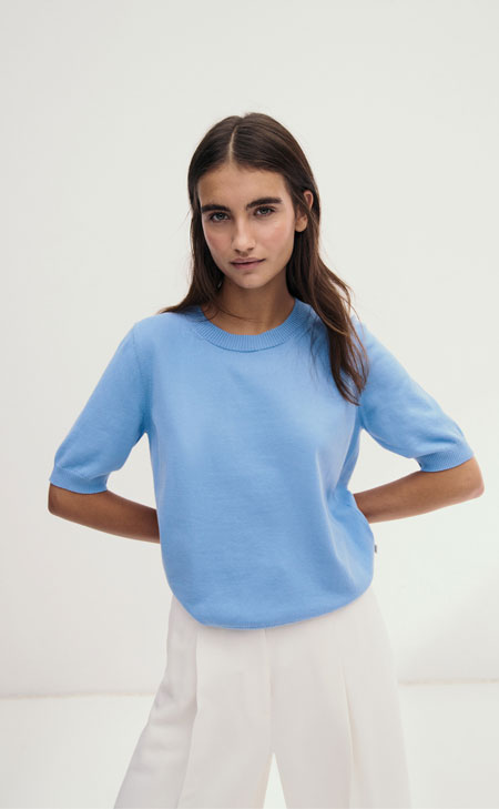 Frau im hellblauen Organic‑Cotton‑Pullover, modern und clean im Studio inszeniert – hochwertige Bio‑Baumwolle für leichte Premium‑Looks.