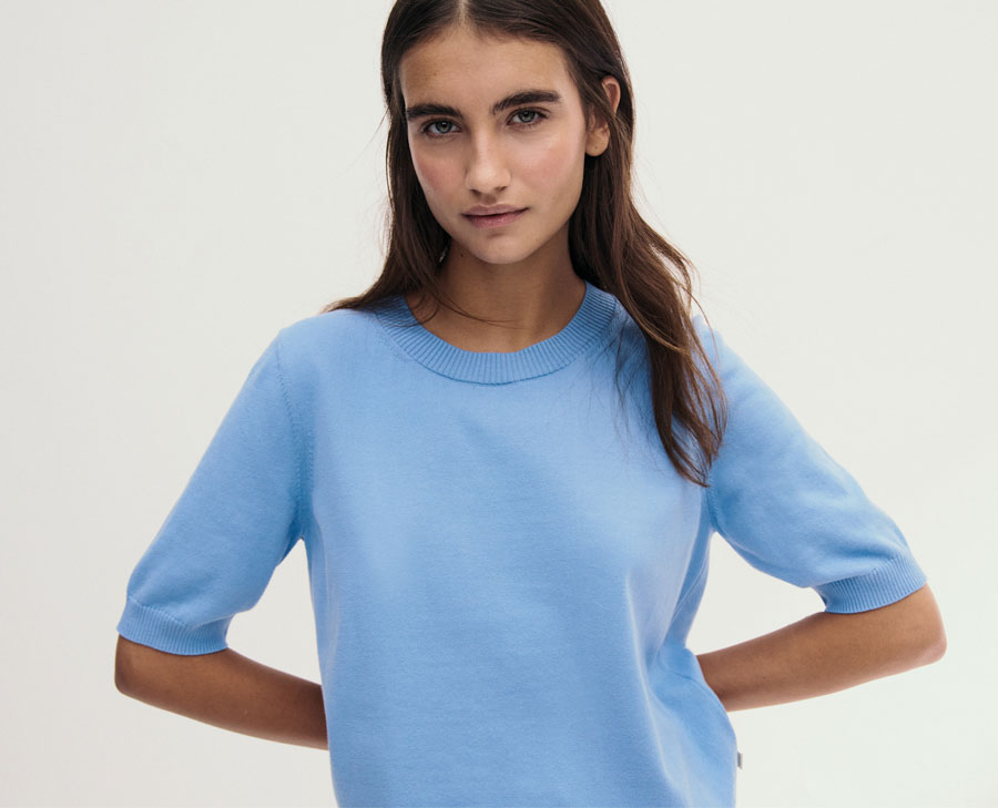 Frau im hellblauen Organic‑Cotton‑Pullover, modern und clean im Studio inszeniert – hochwertige Bio‑Baumwolle für leichte Premium‑Looks.