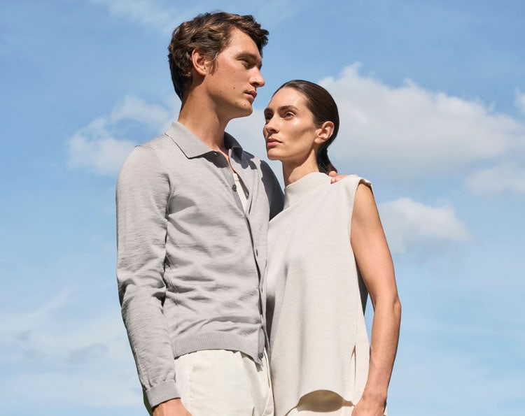 Mann und Frau in leichten Merino‑Air Looks vor blauem Himmel, modern und klar inszeniert – extrafeiner Strick für luftige Premium‑Sommermode.
